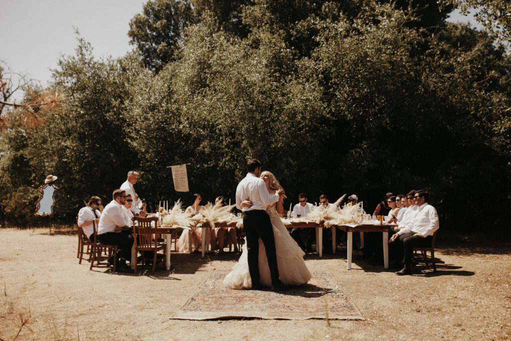 Bride and groom dance in the center of farm tables at their micro wedding in Mariposa, California. Guests seated at the tables watch, with pampas grass, dried florals and taper candles lining the tables.