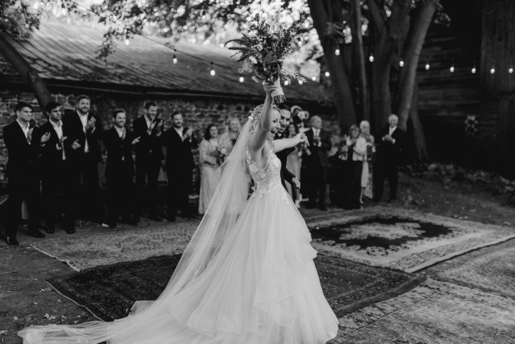 Bride and groom enter their reception space with their wedding party clapping. You can see twinkle lights in the trees and vintage rugs as the dancefloor.