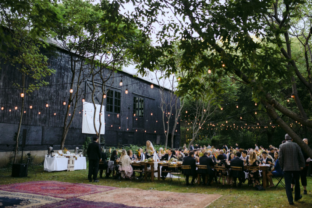 A wedding reception space with guests sitting at long farm tables, a vintage rug dance floor, twinkle lights in the trees, and a black barn at Kester Homestead in the Finger Lakes, NY