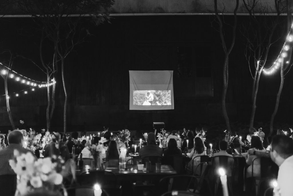 A wedding video plays on a projector at a wedding reception. Guests watch from their seats with twinkle lights and candles lighting the space.