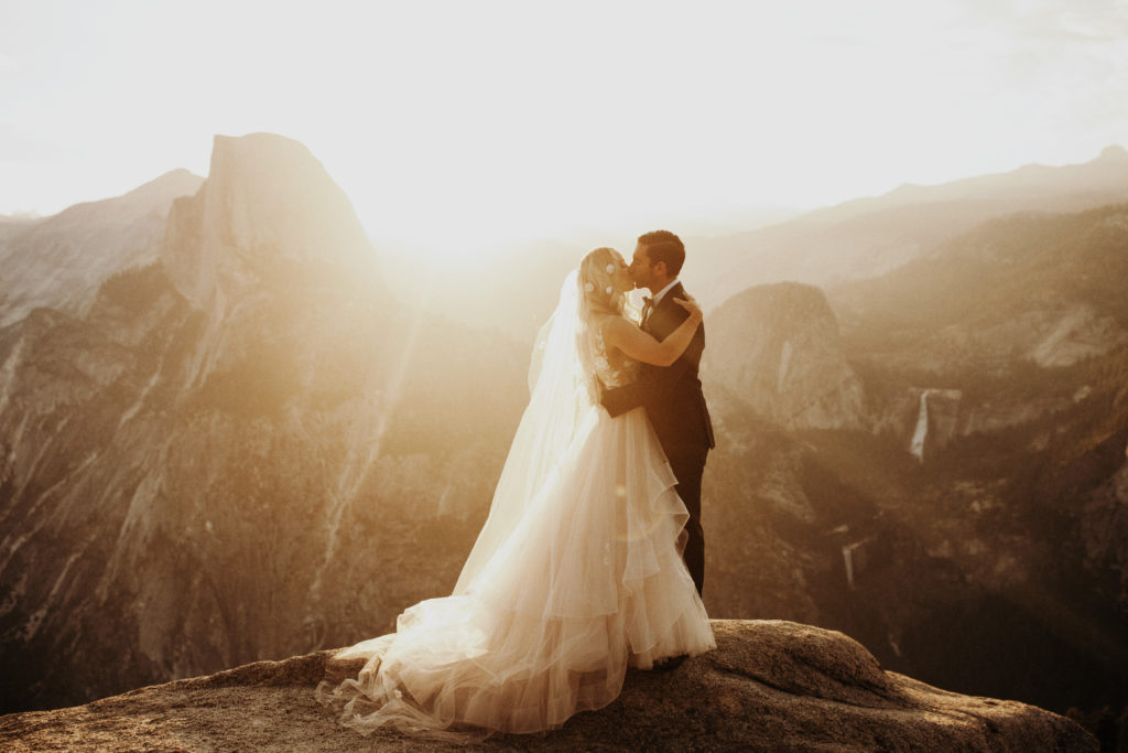Bride and groom kissing on a cliff with the sunrise and half dome behind them. In Yosemite National Park