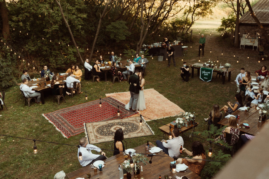 An aerial view of a bride and groom dancing on vintage rugs. Guests surround them, seated at farm tables with string lights hanging above them. Taken at The Kester Homestead, a Finger Lakes wedding venue.