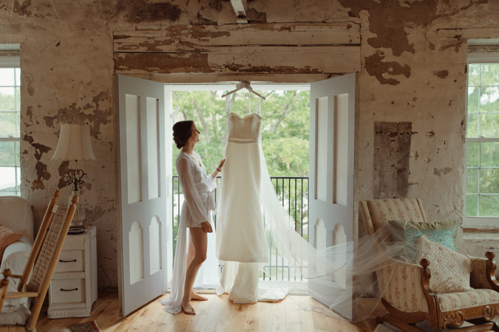 A bride is standing at a window of the Sinclair wedding venue in the Finger Lakes, her dress hanging on the doorframe as she looks at it.