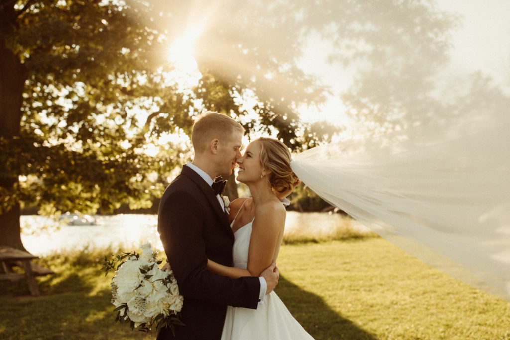 A couple is facing each other on the lawn of the Skaneateles Country Club wedding venue in the Finger Lakes. The bride's veil is blowing behind her as they look at each other, foreheads touching.