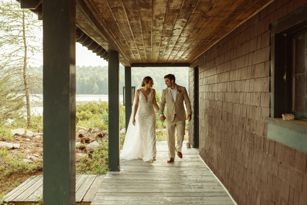 A bride and groom hold hands and walk under a cabin overhang, the bride in a wedding dress and the groom in a tan suit.