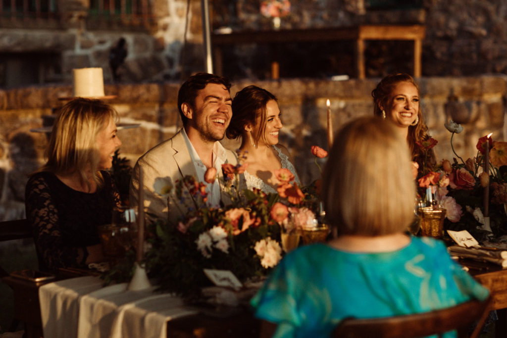 A couple is sitting at their intimate wedding reception. The bride is leaning into the groom, and both are smiling and laughing.