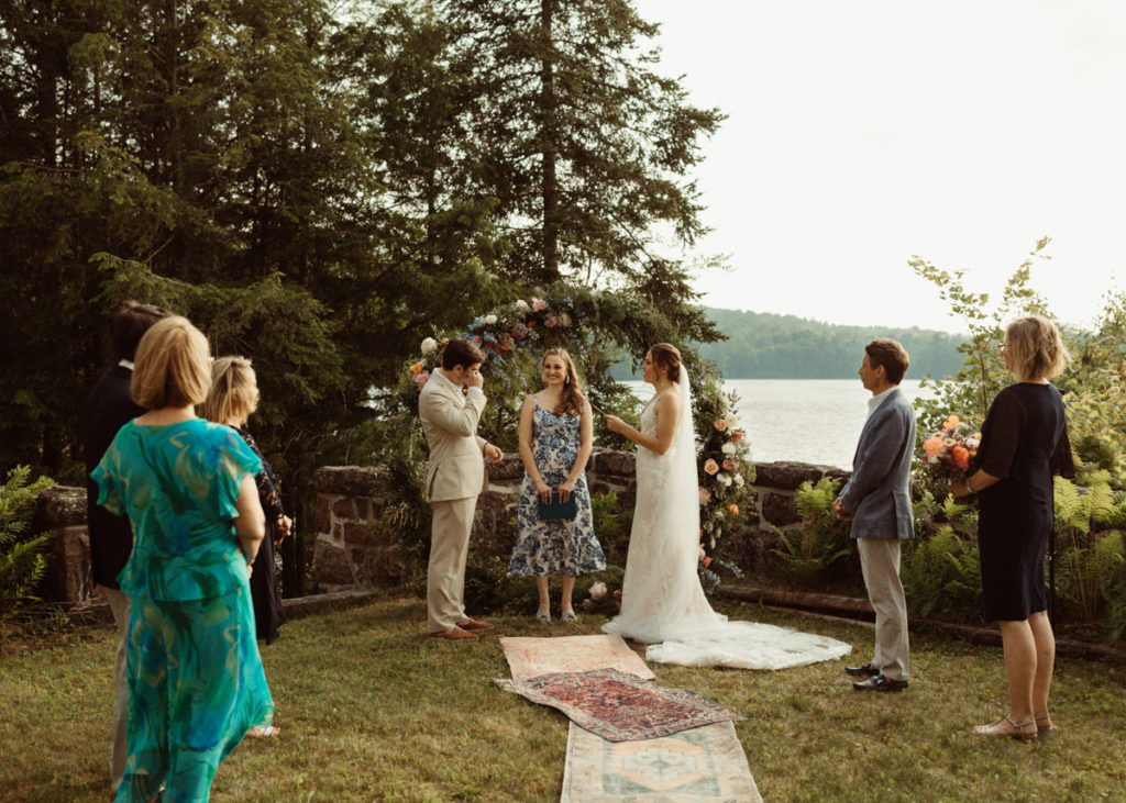 An intimate wedding ceremony is taking place, with a lake in the backdrop. The bride is reading her vows, while the groom stands across from her, wiping a tear.