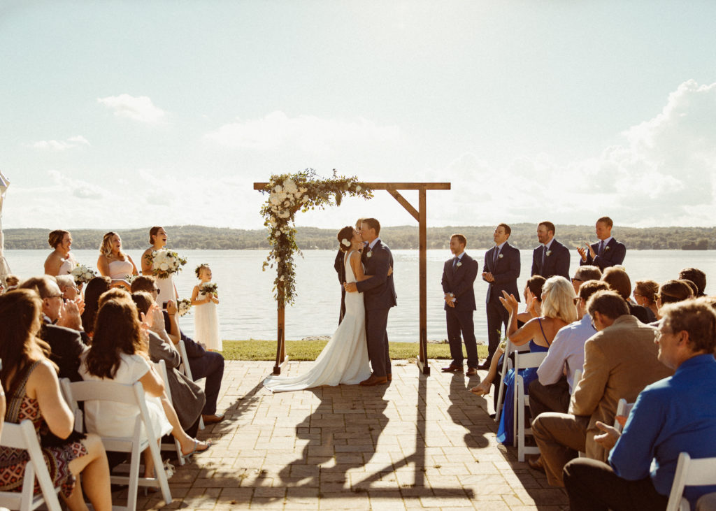 A couple is at the Canandaigua Country Club wedding venue in the Finger Lakes, kissing in front of a lake as wedding guests cheer.