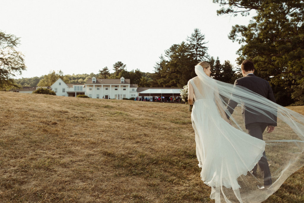 A wedding couple walks up the hill towards their wedding reception. The Fountainebleau Inn is a white manor that sits on a hill with a tent of lights and mingling guests inside. Located in the Finger Lakes of NY.
