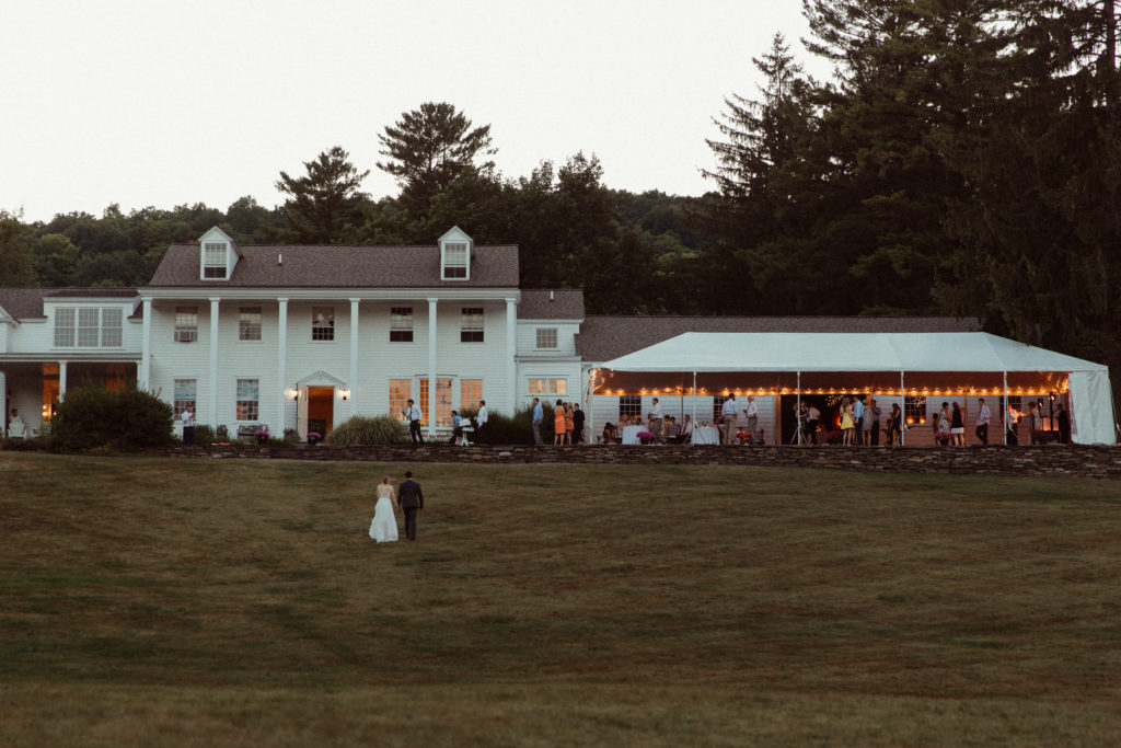 A wedding couple walks up the hill towards their wedding reception. The Fountainebleau Inn is a white manor that sits on a hill with a tent of lights and mingling guests inside. Located in the Finger Lakes of NY.