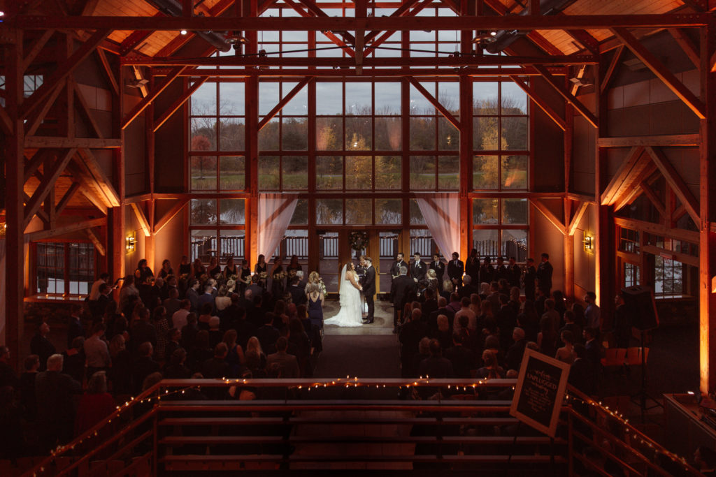 A couple is facing each other during their ceremony at the Lodge Skaneateles wedding venue in the Finger Lakes, with big windows behind them and their guests sitting down.
