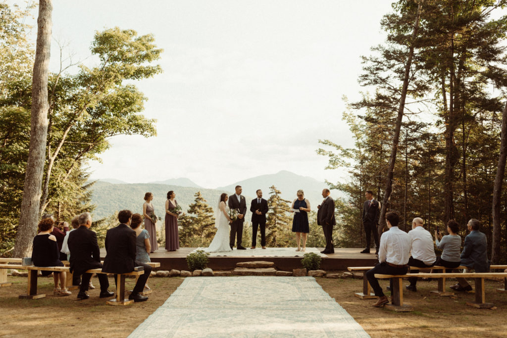 Wedding ceremony takes place overlooking the Adirondack Mountains. The bride, groom, officiant and bridal party stand on an elevated stage with their guests watching below from wooden benches. The trees frame the stage and there are mountains in the distance. At Experience Outdoors in Lake Placid, NY.