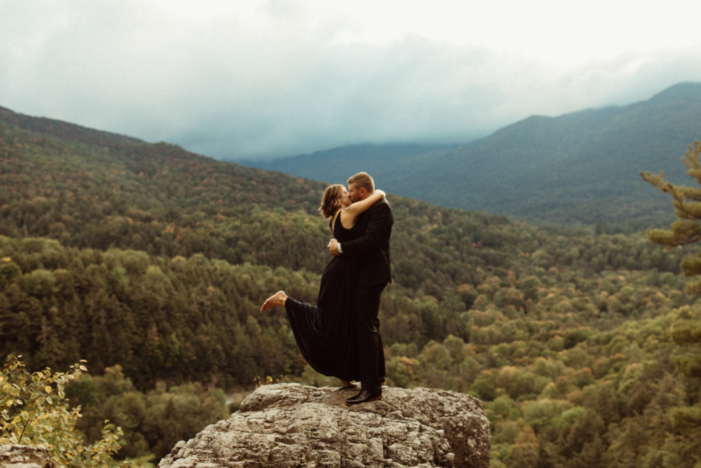 A couple is standing at an overlook in the Adirondacks after their elopement ceremony, and the groom is picking her up by the waist.