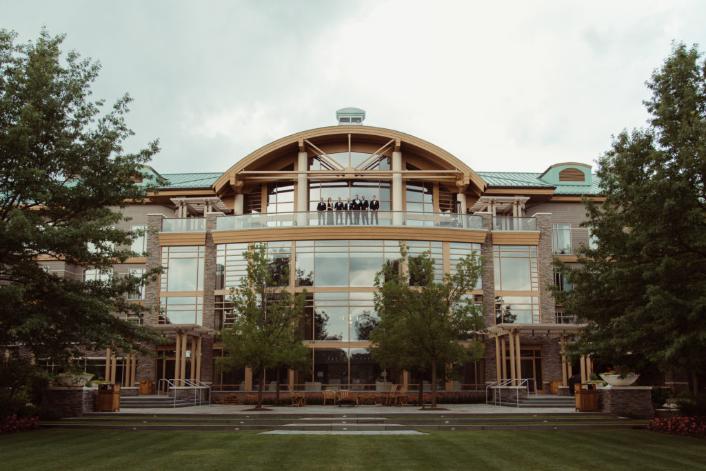 Groom and groomsmen stand on the balcony, overlooking the great lawn at Turningstone Resort and Casino, a wedding venue outside of Syracuse, NY.