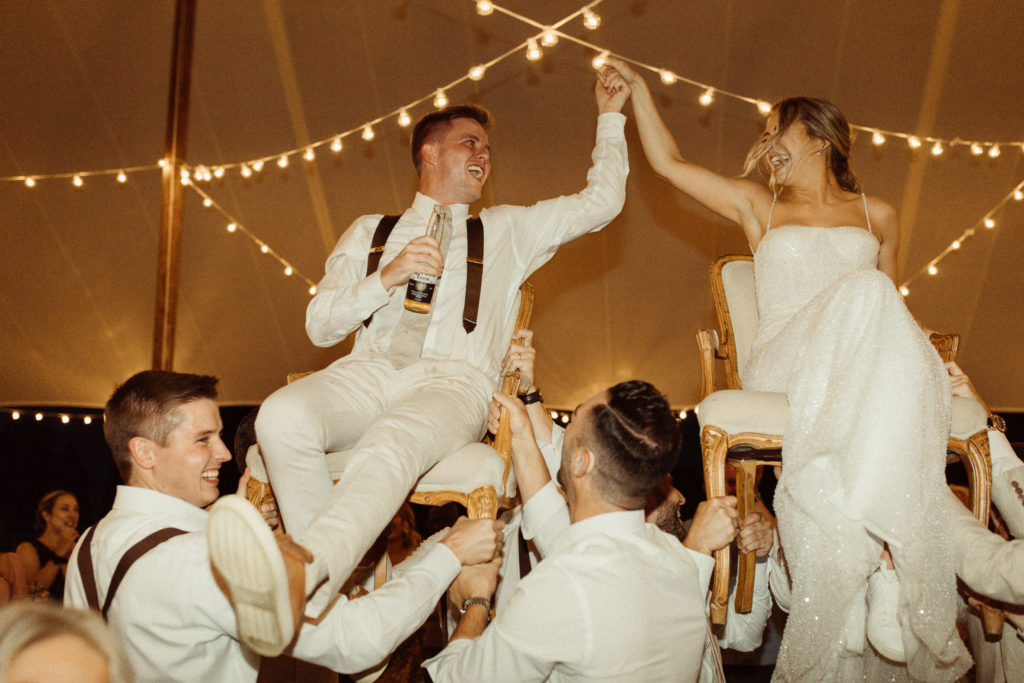 A couple celebrates at their wedding reception. They are hoisted in the air on chairs by their guests, a blending of their cultures and a unique moment at their wedding.