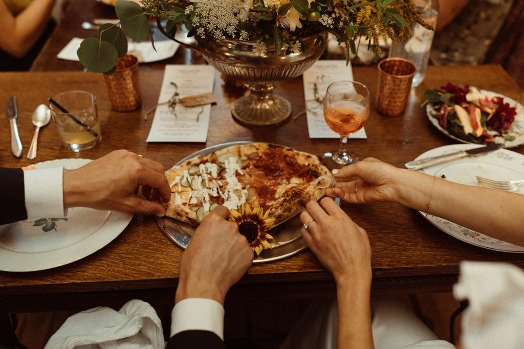 A wedding couple's hands take a slice of pizza at their wedding reception. Showing off their tablescape and signature cocktails which made their wedding unique. Taken at Kester Homestead in Upstate NY.