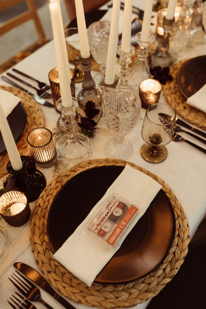 A unique idea for placecards at a wedding reception, cassette tapes with the guests names written in calligraphy. The tablescape is filled with white taper candles and dark accents in the plates and glassware.